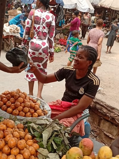 This image captures a vibrant and busy outdoor market scene, likely in a tropical or subtropical region, centered around a vendor interacting with a customer.

In the foreground, to the right, a woman with dark braided hair is seated, looking towards the left with a warm smile. She is wearing a black short-sleeved t-shirt with gold stripes on the sleeves and a white logo on the chest, which appears to be the Real Madrid football club crest. Her right hand is extended, holding paper currency, seemingly in the process of a transaction. On the left, partially visible, another person's hand is holding a black plastic bag and more paper currency, meeting the vendor's hand.

Spread across the bottom of the frame are large quantities of fresh fruit, likely the vendor's merchandise. Two large basins or containers hold piles of small, round, yellowish-orange fruits, which resemble guavas. Mixed among these fruits are fresh green leaves. In the very bottom right corner, a few larger, reddish-yellow fruits, identifiable as mangoes, are also visible.

The background reveals a bustling market environment. Numerous people are walking around, suggesting a busy day. In the mid-ground, a woman wearing a distinctive white dress with red or maroon crown patterns is seen from behind, walking away from the camera. Further into the background, a young child in a green and white striped outfit is visible. Overhead, various tarpaulins or awnings provide shade for the market stalls and pathways. Carts, baskets, and other goods are scattered throughout the background, indicating a diverse array of items being sold. The ground appears to be unpaved, possibly dirt or gravel.

The overall atmosphere suggests an active commercial hub, with the smiling vendor indicating a positive interaction within the busy market setting. The daylight suggests it's daytime.