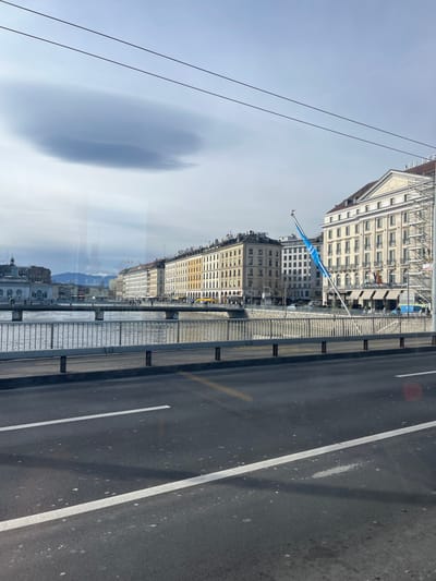 This media file captures a wide urban waterside landscape in Geneva, Switzerland, viewed from a slightly elevated perspective, possibly through a window with significant reflections. The image is taken at daytime under a bright, mostly clear sky with some elongated, wispy clouds.
In the foreground, an asphalt road with white and yellow lane markings is visible, separated by a light-colored barrier or railing from what appears to be a pedestrian walkway or another structure. This suggests the photo might be taken from a bridge or a vehicle on one.
Beyond the railing, a large body of water (Lake Geneva) stretches out, reflecting the sky and distant elements. On the far side of the water, a long row of multi-story buildings with classical architecture lines the waterfront. Many of these buildings feature numerous windows, balconies, and some have awnings or scaffolding. A light blue flag flies from a pole in front of one of the buildings. Distant mountains, possibly the Alps, are visible on the horizon, partially obscured by reflections on the viewing pane.
Notable details include two dark overhead wires cutting across the top left portion of the sky. The most prominent reflection on the viewing surface is a dark, blurred oval shape in the sky area, and a faint reflection of buildings and mountains in the lower half of the image, superimposed over the actual water and land. Text on the scaffolding of one of the buildings is barely legible, possibly indicating "LES BERGUES" or parts of a hotel or cafe name. No explicit people are discernible in the scene.