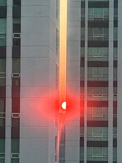 A mid-level, head-on shot of a multi-story building, potentially a hospital or office complex, in daylight. The building's facade features a repetitive pattern of windows, separated by grey panels. A horizontal, orange glow is centered on the image, emanating from behind one of the building's structures, which casts a slight shadow downwards. An area of red light is cast around and below the orange light. The location is potentially Jesús del Monte, Mexico.