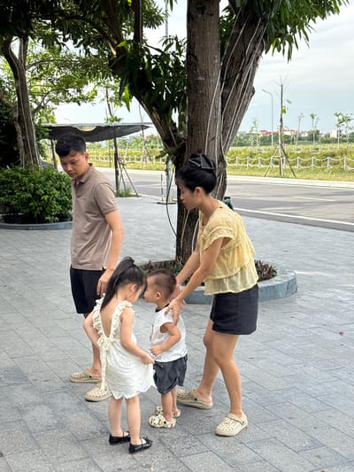 In Hoa Lư Ward, Vietnam, four individuals, two adults and two children, are positioned on a paved area near a street. A man in a tan polo shirt and black shorts stands to the left, looking down. A woman in a yellow ruffled blouse and black shorts is to his right, interacting with the children. The two children, a girl in a white dress and a boy in a white t-shirt and dark shorts, are facing each other. The girl has her arm around the boy. They are all barefoot or wearing casual shoes. The background includes trees, a road, and distant buildings under an overcast sky.