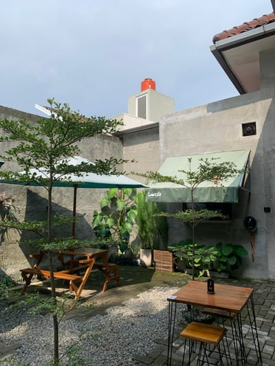 The image displays an unoccupied outdoor patio area of L'e cafe, located in Kota Batu, Indonesia. The ground is primarily covered with light-colored gravel in the foreground, transitioning to dark brick pavers towards the midground and right side. Unplastered, light-grey concrete walls form the perimeter, suggesting an enclosed courtyard space.
On the left side of the patio, a single light-brown wooden picnic table with attached benches is positioned beneath a large, open light-green outdoor umbrella with a white pole. Lush green, large-leafed plants grow along the wall behind this seating arrangement. A slender tree with green foliage stands in the immediate foreground, slightly left of center.
On the right, a small square wooden table with dark brown top and black metal hairpin legs is accompanied by a matching wooden stool tucked underneath. A black table number stand displaying the white numeral '7' rests on the table surface. Above this, a light-green awning with "L'e cafe" inscribed in white lettering extends from the wall, covering a window or service opening. A smaller tree with green leaves is situated directly beneath this awning. Higher on the right wall, a square ventilation fan is visible, with a small black cylindrical object mounted directly below it.
In the background, behind the patio walls, various building structures are visible. On the far right, a section of a building with a brown tiled roof and a gutter is present. Centrally, a light-grey building block rises above the main wall, topped by a prominent red cylindrical water tank against a clear, pale blue sky. Below the water tank, a white, rectangular opening or window is visible on the building block. Shadows from the walls, trees, and umbrellas are cast across the ground, indicating direct sunlight. No individuals are present in the scene.