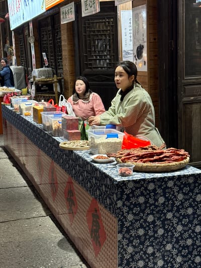 This image depicts a street market stall in Pengshui Miao and Tujia Autonomous County, China, likely during evening hours. Two women are seated behind a long, rectangular display table laden with various food products. The table is covered with a dark blue and white floral-patterned tablecloth, with the lower front section featuring a red checkered design interspersed with large red diamond shapes containing black Chinese characters.
The woman on the right has dark hair pulled back and wears a light green and white striped long-sleeved top, possibly an apron, over a darker garment. Her hands rest on the table near a clear plastic container. The woman on the left, with dark hair, wears a light pink long-sleeved top with a white frilled collar. Both women are looking forward, appearing to attend to the stall. A third woman in a blue jacket is partially visible further down the street to the far left.
The stall displays numerous food items, including various dried goods, nuts, and seeds contained in clear plastic boxes and containers. Several red wooden boxes are also present. On the right, a large quantity of reddish-brown dried meat strips (possibly jerky) is arranged in a flat, round woven basket. Small white dishes and a clear plastic cup hold samples of nuts or dried berries. A flat, round woven tray near the center holds light-colored dried food items, possibly mushrooms or vegetables.
The background consists of dark wooden storefronts with intricate lattice-style windows and doors. Several signs with Chinese characters are visible above and on the buildings, including "饮食有节" and "匠心手." A vertical white banner with Chinese script and an illustration hangs on the wall behind the right woman. The foreground shows grey paving stones on the ground. The scene is illuminated by artificial overhead lighting.