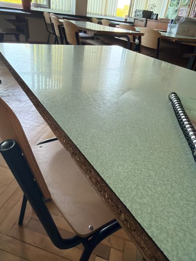 This is a medium-angle, close-up shot of a classroom desk. The desk surface is a light green speckled laminate with visible wear marks. A black spiral-bound notebook is positioned on the right side of the desk. In the foreground, the dark metal legs and beige seat of a chair are visible. The background shows other classroom desks and chairs, a window with vertical blinds, and what appears to be a teacher's desk with electronic equipment. The flooring is a parquet style. The lighting suggests natural daylight entering from the window. There are no people in the frame. The scene is consistent with an empty classroom in Gara Elin Pelin, Bulgaria.