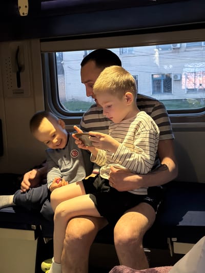 Inside a train carriage, an adult man is seated near a window. The exterior view through the window shows a building and suggests an urban environment. He is holding two young children in his lap. The three are engaged in an activity involving a mobile device. The location information (city of Trudovoi, Russia) is provided, though not visually verifiable from the image.
