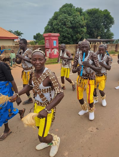 A group of men are seen in Kafanchan B, Nigeria, seemingly performing a traditional dance. The men are dressed in matching outfits consisting of yellow pants with brown stripes, white tank tops, and brown buttoned vests. They are wearing beaded headbands and bracelets, and some have ankle decorations. Several of the men are playing long, blue flutes. The men are standing on an asphalt road with buildings and trees in the background. A red "smartcash" ATM machine is visible in the background. The weather appears overcast.
