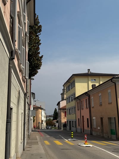 The image depicts a street scene in Balerna, Switzerland, under a clear blue sky. A narrow street is lined with buildings on both sides. On the left, a building with a pink facade and gray shutters is prominent, with a large tree with dark green foliage extending from behind it. The building on the right has facades in yellow, pink, and tan colors, with multiple windows and doors visible. The street is paved with asphalt and features yellow crosswalk markings. Several traffic cones and bollards are placed on the road, some with reflective orange and white markings. In the distance, a car is seen driving down the street. A lone figure, appearing to be a woman in athletic wear, is walking on the sidewalk on the left side of the street. Further down the street, another building with a distinctive bell tower is visible. The overall impression is a quiet, urban environment on a sunny day.