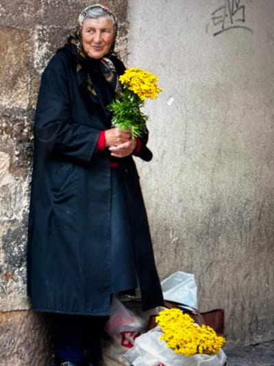 An elderly woman lies on her back on a weathered, uneven ground surface, possibly stone or concrete, with a faint graffiti-like marking on the wall behind her. She is wearing a headscarf and a dark, long coat with red accents visible at the cuffs. She holds a bouquet of yellow flowers and some greenery in her hands, with her eyes closed and a serene expression on her face. To her right, a plastic bag and other items are piled up. The lighting suggests it might be daytime, though the specific time of day and weather are not discernible. The scene appears to be outdoors, possibly in an urban environment given the wall and graffiti, and the general atmosphere is one of rest or peacefulness.