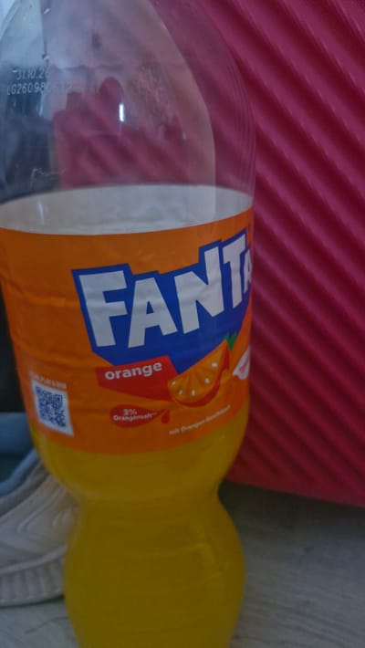 This media file features a close-up, slightly angled shot of a plastic bottle of Fanta Orange, likely a product of Coca-Cola. The bottle is predominantly orange and yellow, with the distinctive Fanta logo in large blue letters on the orange label. The label also displays an image of an orange slice and text that reads "orange" in a red rectangle. Below this, there's text indicating "3% Orangensaft" (3% orange juice) and "mit Orangensaft-Geschmack" (with orange juice flavor). A QR code is also visible on the label.
The bottle is positioned horizontally against a background that appears to be a textured pink surface on the left, with a lighter, possibly wooden or textured gray surface on the right. The lighting suggests an indoor setting. There are some reflections on the plastic bottle, indicating it's likely a clear bottle with the beverage inside.
Visible text includes the brand name "FANTA," "orange," "3% Orangensaft," "mit Orangensaft-Geschmack," and what appears to be a batch code or expiration date printed on the clear part of the bottle: "-31.10.26 LG260980632".
The context provided indicates the location as Uithuizen, Netherlands. No people are visible in the image, and there are no strong cues about the time of day or weather. The overall impression is a product shot, focusing on the branding and details of the beverage bottle.