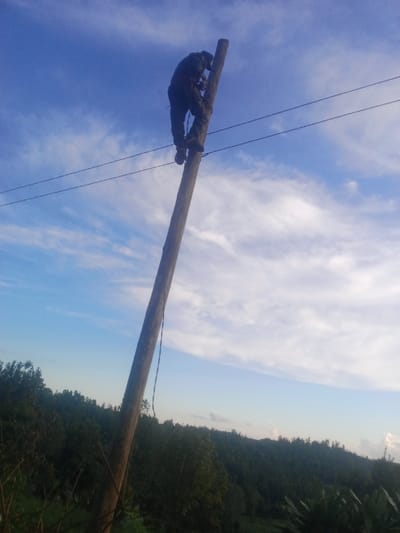 A solitary individual is depicted ascending a wooden utility pole in Karuri, Kenya. The person, dressed in dark work attire and utilizing climbing gaffs affixed to their boots, is positioned against the pole approximately two-thirds of the way up. Multiple dark utility wires are visible extending horizontally from the pole near the worker's location. A dark rope or strap hangs down the pole from the individual. The sky overhead is blue with scattered white clouds, while the background reveals a verdant, hilly landscape densely covered in green trees and vegetation.