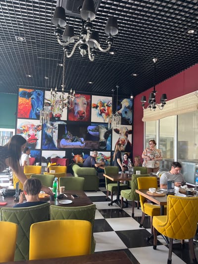 The image depicts the interior of a restaurant, likely located in Akhlebaikha (Kovriginskii S/S), Russia, characterized by its modern and artistic decor. The dominant feature of the ceiling is a black grid pattern with recessed lighting and two ornate chandeliers. The walls are adorned with a vibrant collage of large paintings, predominantly featuring stylized portraits of cows in various colors and expressive poses.

The restaurant's seating arrangement includes tables with dark wood tops and chairs upholstered in bright yellow and deep green velvet, many with decorative nailhead trim. The floor is a striking black and white checkerboard pattern.

Several individuals are visible within the restaurant. In the foreground, a young person with short brown hair is seated at a table, looking at their phone while a pizza sits before them. To their left, a woman with long dark hair, seen from the back, appears to be interacting with the young person or the table. In the middle ground, a group of people are seated at a table, including a man in a grey t-shirt looking at his phone, and a woman with sunglasses and a floral-patterned shirt holding a purse. Further back, other patrons are seated at tables and conversing. The overall atmosphere suggests a casual dining experience.