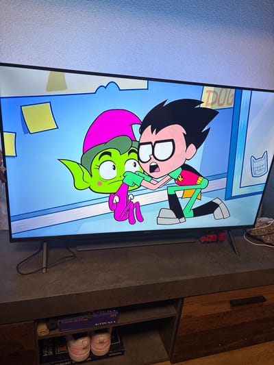A flat-screen Philips television, displaying an animated scene from "Teen Titans Go!", dominates the center of the image. The screen shows two cartoon characters: one with green skin, large ears, a pink pointed hat, and a pink bodysuit (Beast Boy), appearing wide-eyed and surprised. The other character, with black hair, glasses, and a red, green, and yellow superhero suit (Robin), is kneeling, with both hands extended towards the face of the green character. The on-screen background is a light blue wall adorned with several yellow sticky notes and a partially visible sign that reads "DUG" in yellow letters. To the far right of the screen, a stylized angry cat emblem is visible on a light blue door frame.
The television rests on a dark gray media console with a textured surface. On the console, to the right of the television's center, are two small red toy vehicles, a truck and a trailer. Black electrical cables are visible behind the television and console. To the right of the console, a white power cable is coiled next to a small, textured, brown, cylindrical object.
Below the console, two open shelves are visible. The top shelf contains a purple box labeled "VIVALUX LED DOLCE/R LED 15W BK CL" and two small, brown, textured circular objects. The bottom shelf holds a pair of pink and white Nike sneakers, positioned atop a partially visible box, likely a "500 PIECE PUZZLE", and a box or book labeled "HAWAII SPACE". To the right, a wooden drawer with a black rectangular handle is integrated into the console. The floor beneath the console is light brown wood. The wall above the television has a light, textured finish. This scene is located in a residence in Kuklen, Bulgaria.