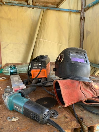 The image shows a collection of welding and grinding equipment, likely in a workshop or outdoor industrial setting. The primary objects include a black welding helmet with a purple tinted visor, a pair of reddish-brown welding gloves hanging on the helmet, an orange and black portable welding machine with a digital display, a clear plastic bottle of water, and a teal and black angle grinder. The equipment is placed on a rusty metal surface, suggesting a work area that might be exposed to the elements. The background consists of a yellowish tarp or canvas material supported by a weathered blue and rust-colored metal frame. There is no visible indication of the time of day or weather conditions beyond the implied outdoor setting.

Visible text includes:
- On the welding machine: ".Sturm!" and "AW971550"
- On the bottle label: Part of a label with Russian or Ukrainian text is visible, including "ТЕ"
- On the angle grinder: A label with text and a model number is partially visible, including "15328-PE" and a Japanese industrial standard symbol.
- On a box next to the water bottle: "MAXweld" is clearly visible.

The overall impression is one of tools and gear prepared for or recently used for metalworking tasks.