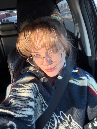 A female individual is seated inside a vehicle, occupying what appears to be the driver's or front passenger's seat, indicated by the diagonal seatbelt worn across her chest and shoulder. She has light-colored hair with bangs, pulled back into a partial bun secured by a light green hair tie. Her eyes are light blue, and she wears rectangular eyeglasses with metallic frames. She is dressed in a dark, possibly brown, high-neck garment underneath a patterned jacket featuring a dark blue base with prominent white, red, and yellow abstract designs. Her gaze is directed towards the camera, and her lips are closed in a slight smile. Strong natural light illuminates her left side, casting highlights on her hair and face. The car's interior features black leather seating with a quilted headrest. Through the car windows, a snowy or icy exterior environment is visible, along with distant structures suggesting a residential or rural setting. The scene is located in Азьмушкино, Russia.