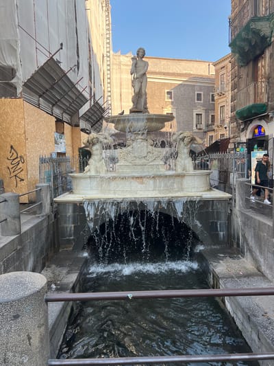 The image shows a public fountain in an urban setting, likely Catania, Italy, given the context. The fountain is ornate, made of white stone, and features sculptures of mythological figures, with water actively flowing and splashing. The water cascades into a dark basin, with visible ripples and spray.
In the background, buildings are visible. One building on the left is covered in scaffolding and white tarpaulin, suggesting ongoing renovation or construction. Another building on the right appears older, with a more traditional architectural style. There are people present, one standing near the top of the fountain and another seated on a ledge. The scene is lit by natural sunlight, indicating it's daytime. The sky is clear and blue.
The perspective of the image is slightly tilted, giving a dynamic feel to the scene. The overall impression is of a lively public space with historical elements. There is some graffiti visible on a wooden panel in the foreground, with the word "DUNK" clearly legible. A neon sign with blue and yellow elements is also visible in the upper right portion of the image.