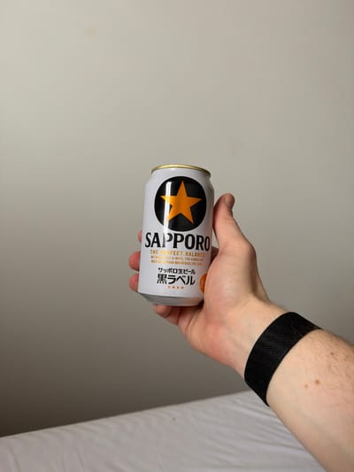 A person's hand holds a white Sapporo beer can with a gold top. The can's label features a black circle with a gold star in the center and the word "Sapporo" in black lettering. Below, text in English and Japanese provides details about the beer. The person wears a black band around their wrist. The background is a plain white wall on the left and white sheets on the right. The image is rotated counterclockwise, causing the objects to appear angled.