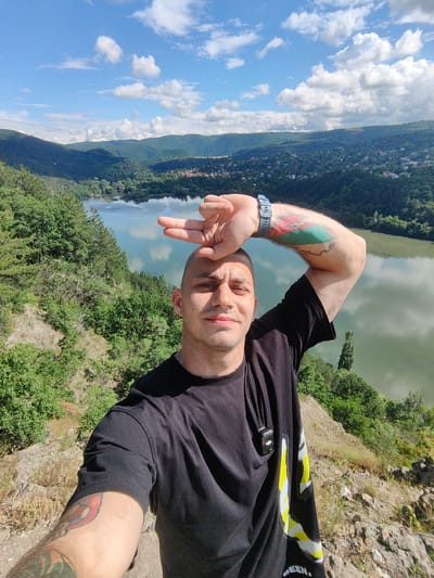 This is a selfie taken by a man standing on a rocky overlook, with a scenic view of a river valley under a bright, cloudy sky. The man, who appears to be in his late 20s or early 30s, has a shaved head and is wearing a black t-shirt. He is posing for the photo with his right arm raised, holding two fingers to his forehead. He has tattoos on both arms, with a visible tattoo on his right bicep featuring green, red, and white colors, possibly representing a flag. A wristwatch is also visible on his right wrist. He has a small black device attached to his t-shirt near his collarbone.
The background of the image showcases a lush, green landscape. A wide river or lake is visible in the mid-ground, reflecting the sky and surrounding trees. The valley is densely covered with trees, and a town or village is nestled on the hillsides in the distance. The terrain appears hilly and somewhat rugged.
The time of day appears to be daytime, with bright sunlight casting shadows. The weather seems pleasant, with scattered white clouds against a clear blue sky, suggesting a warm day. The location appears to be a natural, possibly mountainous or hilly, area. The man's pose and the selfie format suggest a casual, perhaps recreational activity like hiking or sightseeing. The overall mood of the image is relaxed and happy, with the man appearing content and enjoying the view. There is no visible text in the image.