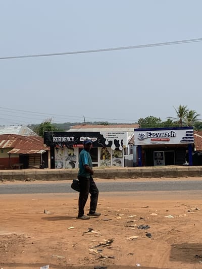 The image depicts an outdoor scene in Ilorin, Nigeria, along a street. A man wearing a blue shirt and dark pants is walking along the right side of the road. To the left of the road are commercial establishments. A business called "RESIDENCY" with signage indicating "WIG INSTALLATION, BARBING, COSMETICS," and another named "Easywash LAUNDRY SERVICE" are visible. A utility pole with wires is seen against a bright sky. Debris is scattered on the unpaved ground beside the road.  A palm tree is present near the businesses. Adjacent to the commercial buildings is what looks like the edge of residential housing with tin roofs.
