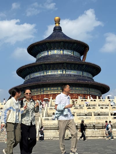 The image depicts the Hall of Prayer for Good Harvests, a key structure within the Temple of Heaven complex in Dongcheng District, China. The iconic multi-tiered, circular building with a dark blue, tiled roof and a golden finial is centrally located. In the foreground, three individuals are walking. The two women on the left are in focus, with one wearing a light-colored jacket and cargo pants, and the other in a floral-patterned shirt and dark pants. Both appear to be holding snacks. A man in a white polo shirt and beige pants is walking in front of them, to the right. Behind these individuals, a wide stone staircase leads up to a terrace where more people are visible, some holding umbrellas. The sky is blue with scattered clouds, suggesting a sunny day. The overall scene captures a moment of tourism and exploration at this significant historical site.