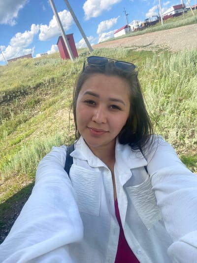 A woman takes a selfie outdoors with a red outhouse and buildings in the background. She is wearing a white button-down shirt and sunglasses are on top of her head. The landscape is grassy with a dirt road. The sky is blue with white clouds.