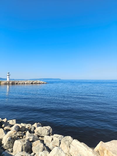 The image shows a bright, clear day at the coast. In the distance, on a rocky outcrop, stands a white lighthouse with a green top. The lighthouse is reflected in the calm blue water. The foreground is filled with large, light-colored rocks. The vast expanse of the sea stretches out to the horizon, meeting a clear blue sky. There are no people visible in the image. The location is identified as Volzhskiy Utes, Russia.
