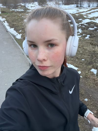 This is a vertical selfie of a young woman outdoors in Berezniki, Russia, likely during late winter or early spring. The subject is a Caucasian woman with light skin, numerous reddish freckles across her cheeks and nose, and light green eyes. Her light brown hair is pulled back, and she is looking directly at the camera with a neutral to slightly smiling expression.

She is wearing white over-ear headphones and a black athletic jacket with a prominent white Nike swoosh logo on the left chest. A white smartwatch or fitness tracker is visible on her left wrist.

The setting is an outdoor area featuring patches of melting snow scattered on dry, brown grass or ground, indicating a cool, transitional season. Bare trees are visible in the background, further supporting the late winter or early spring timeframe. A grey concrete path or sidewalk runs along the lower and right portions of the image, suggesting she is on a pathway. The lighting suggests it is daytime. The overall scene depicts an individual engaged in an outdoor activity, possibly a walk or run.