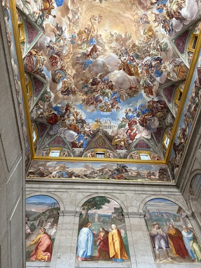 The image depicts a grand, ornate interior space within the Royal Monastery of San Lorenzo de El Escorial, San Lorenzo de El Escorial, Spain. The scene is dominated by extensive fresco painting covering the vaulted ceiling and upper walls, with additional murals on the lower wall sections.

The vaulted ceiling is covered by a vast Baroque fresco, portraying a dynamic celestial scene. Hundreds of human and angelic figures are depicted in various active poses, ascending through swirling clouds towards a central, luminous light source. Elements such as a cross are visible being carried aloft. The color palette is vibrant, featuring prominent blues, whites, reds, greens, and gold accents.

Below the main ceiling fresco, the upper wall sections feature smaller, horizontally oriented rectangular murals. These murals depict historical or battle scenes, including numerous human figures, cavalry, and detailed landscapes, rendered in a more subdued, earthy color scheme. These painted sections are framed by ornate architectural elements and gold trim. Several small, rectangular windows are visible within these upper painted areas, allowing natural light.

The lower section of the wall is constructed from large, visible cut stone blocks. Embedded within this stonework are three distinct, large arched murals. The leftmost mural, partially visible, shows a cloaked figure kneeling before another against a landscape background. The central mural depicts a standing figure in a blue robe conversing with two cloaked figures, one in red and one in gold, beneath a tree with distant architectural elements. The rightmost mural presents a group of at least four standing figures in various cloaks and robes, engaged in interaction within a landscape that includes a distant city.

The overall perspective is upwards, emphasizing the immense scale and intricate detail of the integrated ceiling and wall artistry.