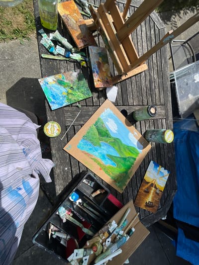 The image depicts an outdoor painting setup on a wooden table in Maidstone, United Kingdom. An easel sits at the table's edge, alongside a palette with dried paint. Several partially completed paintings are scattered on the table, depicting landscapes and buildings. Two jars with lids and tubes of paint are also present. A black container holds an assortment of paintbrushes and painting tools. A person wearing a paint-splattered shirt is positioned in front of the table. The setting includes a partial view of pavement and lawn in Maidstone, United Kingdom.