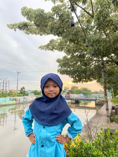A young child, presumably female, stands centrally in the foreground, facing the camera with hands on hips. She wears a dark blue hijab with pleated detail and light blue zig-zag stitching, paired with a long-sleeved, light blue tunic-style dress featuring dark blue buttons and matching zig-zag stitching.

The background depicts a waterside environment in what is identified as Kalitengah, Indonesia. To the child's left, a canal or narrow river flows, its far bank adorned with a low wall painted with a colorful mural. In the mid-ground, a light blue concrete bridge with yellow and red railings spans the waterway; a person on a pink scooter is visible crossing the bridge from right to left. Beyond the canal and bridge, various trees and residential structures are discernible, along with power lines and utility poles. The sky is partially cloudy, showing an orange and yellow glow near the horizon on the right, indicative of dawn or dusk. Overhead, a large tree with variegated green and light-colored leaves partially shades the scene, with a string of dark-colored, bell-shaped lights suspended from its branches. A concrete pathway runs along the canal bank to the child's right, bordered by green foliage and some yellow-flowering plants.