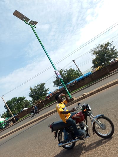 The image captures a street scene in Garko, Nigeria, on a clear day with a few clouds in the sky. A tall, green lamppost with two solar panels is a prominent feature, extending upwards and slightly to the left of the frame. A man wearing a yellow t-shirt with blue accents and blue jeans is riding a red motorcycle in the foreground, moving to the right. He is looking up and to the left, with his right hand raised as if shielding his eyes from the sun. The motorcycle has chrome details and spoked wheels. In the background, another motorcycle with two riders is visible further down the road, heading in the same direction. The street is paved, with a concrete curb and a sidewalk. Beyond the sidewalk, there are buildings and trees. Some of the buildings are behind a reddish-brown wall, and a few have blue tarpaulins covering parts of them. There are also power lines stretching across the frame. To the left of the lamppost, a green truck and a small stand or kiosk are partially visible. The overall impression is of a typical day in an urban environment, with ongoing traffic and street activity.