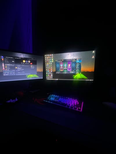 The image depicts a dimly lit room with two computer monitors and a keyboard. The left monitor displays a Discord interface with chat messages and user avatars. The right monitor shows a League of Legends champion select screen with various character portraits and an animated background of a starry night sky over a green tent. A keyboard with RGB backlighting, showing purple and blue hues, is positioned in front of the monitors. Various small, white objects, possibly cable ties or earbud cases, are scattered on the desk to the left of the keyboard. A red gaming mousepad is partially visible under the keyboard. The background consists of a dark wall, with a subtle blue light casting a shadow on the left side of the frame. The overall scene suggests a gaming setup in a private residence, likely a bedroom or dedicated entertainment space. The location in the city of Kazanlak, Bulgaria is not discernible from the visual information provided in the image.