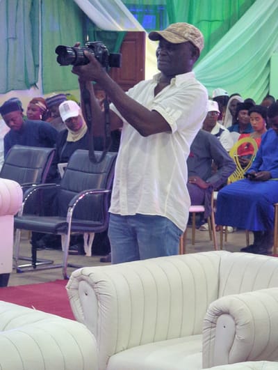 In Ungwan Liman, Nigeria, a man wearing a tan cap, a white short-sleeved shirt, and blue jeans is holding a camera, possibly taking a photograph. The environment appears to be indoors, with green drapes visible on the left. Several other people are seated in the background, including a person in a blue garment with a yellow, hoop-shaped object. Stacks of white, rolled-up materials are on the right, while office-style chairs are visible in the middle ground, occupied by people who may be observing the photographer.