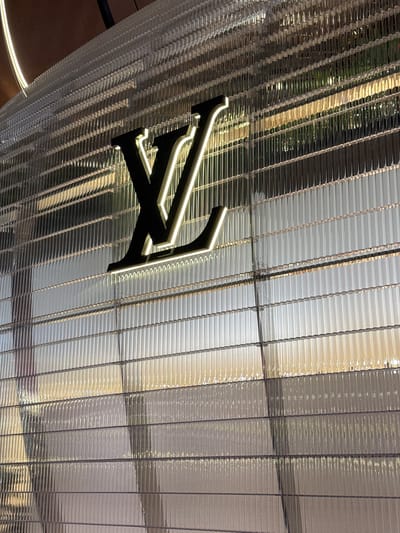 The image shows a close-up of the Louis Vuitton storefront in Doha, Qatar. The facade of the building is covered in a translucent, ribbed material that creates a textured effect. A large, illuminated Louis Vuitton logo is prominently displayed on the facade. The lighting suggests it is nighttime or dusk, with interior lights glowing from behind the facade and illuminating the logo. The overall impression is one of modern luxury and sophisticated branding.