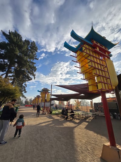 The image shows a sunny day in the city of Puente Alto, Chile, likely in a park or outdoor area. In the foreground, there is a row of brightly colored, Asian-style architectural elements, possibly lanterns or decorative structures. These structures are predominantly yellow, red, and blue. The image is taken from a low angle, emphasizing the height of these structures against a backdrop of a partly cloudy sky and a tree. People are walking along a dirt path. There is a seating area with people at tables.
