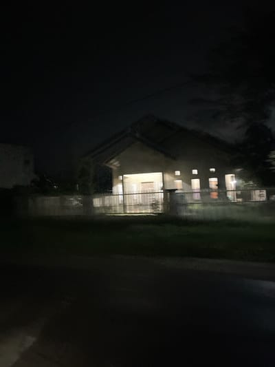 The image shows a house at night. The house is light brown with a dark roof. Several windows are illuminated, suggesting occupancy. There is a fence in front of the house, and trees are visible in the background. The foreground is dark and appears to be a grassy area. The image is grainy due to low light conditions. The location is identified as Landuh, Indonesia.