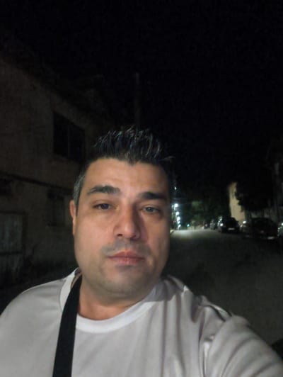 A man with dark, spiky hair and a light complexion is captured in a close-up, selfie-style photograph taken at night in Harmanli, Bulgaria. He is wearing a white t-shirt with a black strap visible across his chest. The background is dimly lit, showing the facade of a building on the left and a street scene with parked cars and streetlights on the right. The overall impression is of a casual, nighttime snapshot in an urban environment.