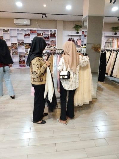 The image shows a shop interior in a clothing store located at the coordinates -6.763, 111.03 in Kecamatan Margorejo, Indonesia. Two women are shopping for clothes. The woman on the left is wearing a black headscarf and a brown sweater with "GG" patterns. She is holding a white scarf in her left hand and is looking at the clothes displayed on a rack. The woman on the right is wearing a brown headscarf and a white blouse with brown patterns. She is wearing a white handbag on her left shoulder and is looking at the clothes displayed on the rack. The clothes on the rack are mostly white and pink. There is a large white skirt hanging on the rack behind her. Both women are wearing black pants. The floor is tiled with large rectangular tiles. The room is lit by overhead fluorescent lights. The background shows more clothes racks with a variety of garments. There are shelves on the left wall with various items. There is a mirror on the right wall showing a reflection of the shop interior. The image is taken at eye level.