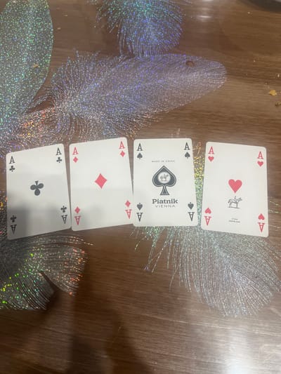 Four playing cards, all Aces, are laid out on a wooden table. From left to right, the cards are the Ace of Clubs, the Ace of Diamonds, the Ace of Spades, and the Ace of Hearts. The Ace of Spades card has "Piatnik VIENNA" and "MADE IN CHINA" printed on it, along with a logo of a horse and rider within a spade. The Ace of Hearts card has a similar horse and rider logo within a heart, and the text "17.310 piatnik.com". The table is decorated with shimmering, feather-like ornaments. There are no people or animals visible in the image. The location of Rosenovo, Bulgaria is not depicted or suggested by the image content.