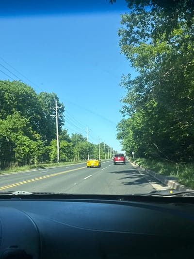 The image depicts a view from inside a vehicle, looking forward onto a road with two lanes separated by double yellow lines. The road is lined with green trees on both sides. Two vehicles are visible ahead: a yellow car closer to the camera and a red car farther away. Overhead power lines are present. The sky is blue and clear. The location may be Morton Grove, United States of America.