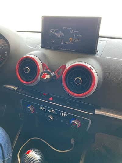 The image shows the interior of a car dashboard with a central infotainment screen displaying car settings. The screen shows options like "Vehicle", "Sound", "Radio", "Media", and "Telephone". A graphic of a car is also visible. Below the screen are two circular air vents with red trim, a hazard light button, and climate control dials and displays showing a temperature of 19.5°C. A USB cable is plugged into the dashboard. The car appears to be stationary. There is no visible indication of the location being in Boughezoul, Algeria.