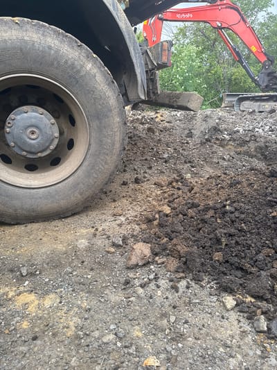 This image depicts an outdoor construction or excavation site in Konstantinovo, Bulgaria, during daytime. The scene is dominated by heavy machinery and disturbed earth.
In the foreground, a large, weathered tire, likely from a truck or heavy equipment, occupies the lower left and center. Visible text on the tire includes "MM 228", "ALL STEEL RADIAL", "315/80 R 22.5", and "RETREAD MADE IN BULGARIA," clearly indicating its manufacturing origin. The tire is dirty, with mud and soil adhering to its surface and rim.
To the left and upper-left, part of a red Kubota excavator is visible. The brand name "Kubota" is prominently displayed on its arm, along with a yellow warning symbol. The excavator's arm and a portion of its bucket or attachment are extended, suggesting ongoing or recent earthmoving operations.
The majority of the background and right side of the image consists of excavated ground. This area features a mixture of dark, rich soil, potentially topsoil, alongside lighter, gravelly earth and scattered rocks. The ground is uneven and appears freshly disturbed. In the upper background, green foliage from trees is visible, indicating a natural or rural setting beyond the immediate work area. The lighting suggests an overcast day or indirect sunlight, providing diffused illumination across the scene. The overall impression is that of an active work site for earthworks or infrastructure development.