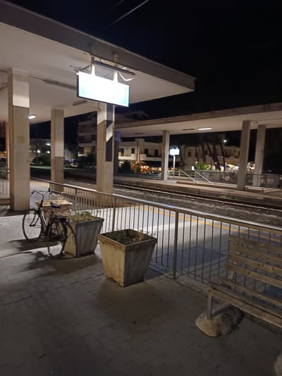 This is a night-time, eye-level, medium-shot photograph taken at the Roseto degli Abruzzi train station in Italy. The scene depicts a train platform with tracks visible in the background. On the platform, there is a bicycle parked next to two large planters. A wooden bench is situated to the right of the frame, and a metal railing separates the platform from the train tracks. The platform is paved with interlocking bricks. Overhead, a canopy supported by pillars provides shelter. A lit digital display board is mounted above the canopy. The surrounding area shows buildings and trees, illuminated by artificial lights, suggesting an urban or semi-urban setting. The image is characterized by low light conditions typical of nighttime, with artificial lighting casting shadows. There are no people visible in the frame.