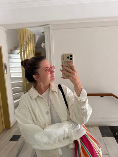 A young woman stands in what appears to be an apartment building hallway or entrance area in Glifada, Greece. She is holding a smartphone up to take a selfie or a photo. The camera points toward a stairwell leading downwards. The stairwell has brass-colored railings and white steps. She is wearing a white jacket with embroidery, a grey shirt, and pink-framed sunglasses. A colorful skirt or pants is visible in the upper right. The hallway is tiled with black and white marble-like material. A door frame is visible above the woman and a light fixture in the stairwell.
