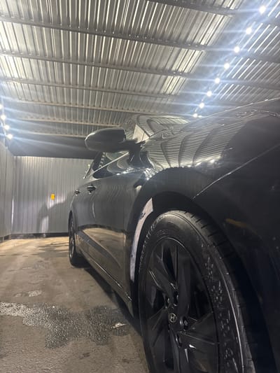 This is a close-up, sideways shot of a black car parked inside a structure with a corrugated metal wall and ceiling. The car's wheel and lower body panels are prominently visible. The interior of the structure is illuminated by a series of bright, overhead lights, casting strong reflections on the car's polished surface. The floor appears to be wet and speckled with debris. A portion of a black side mirror is visible on the car. The image conveys a sense of a car being kept in a covered, possibly industrial or workshop-like, environment. The lighting suggests it could be nighttime or the interior is designed to be brightly lit regardless of external conditions. There is no visible text in the image.
