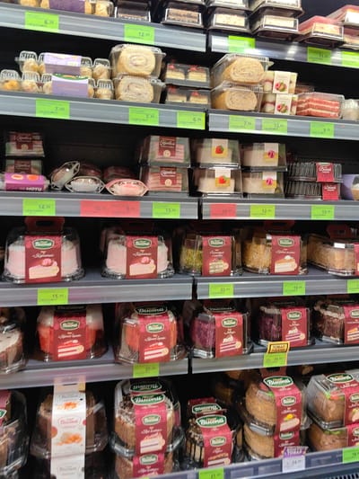 This is a medium shot, eye-level photograph of a grocery store aisle displaying a variety of cakes and pastries on shelves. The items are packaged in clear plastic containers with colorful labels, predominantly featuring the brand "Bisconti". The cakes range in flavor and appearance, including roulades, layered cakes, and tarts, with visible ingredients like cream, fruit, and chocolate. Price tags are visible on some shelves, indicating sales or prices in local currency. The setting appears to be a well-lit supermarket aisle, typical of a European setting, given the location context of Fanipal, Belarus. The image focuses solely on the products, with no people visible. The overall impression is one of commercial display and consumer goods.