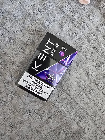 A pack of Kent Sticks with "Berry Click" flavor is placed diagonally on a textured gray blanket. The pack is black with white and purple branding. The text on the pack is partially visible, including the brand name "KENT STICKS" and the flavor "BERRY CLICK". Below the brand name, there is a purple geometric design. On the lower portion of the pack, there is a white label with Russian text, which translates to "SMOKING TOBACCO IS HARMFUL TO YOUR HEALTH". The lighting is even, and the focus is on the product. The scene is static and appears to be a product shot. There is no indication of the location in Yoshkar-Ola, Russia.
