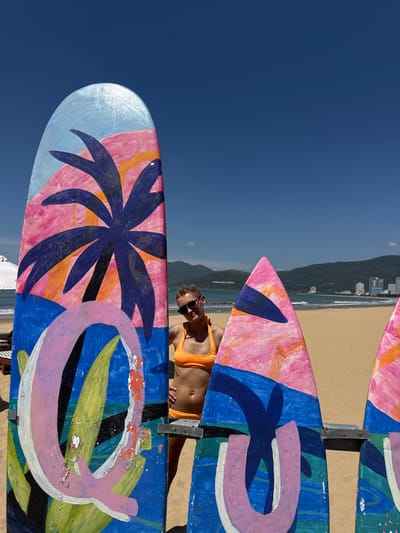 A person in an orange bikini stands behind two colorful surfboards on a sandy beach. The surfboards are decorated with abstract art featuring palm trees, tropical foliage, and geometric shapes in shades of blue, pink, orange, and green. The person, wearing sunglasses, is facing the camera with a slight smile and has short blonde hair. The ocean is visible in the background with gentle waves, and beyond that, a coastline with mountains and buildings. The sky is clear and blue. The scene appears to be in Quy Nhon Ward, Vietnam, given the presence of the ocean, beach, and mountainous landscape characteristic of the area. The surfboards are mounted on metal stands, suggesting they might be part of a display or art installation on the beach.