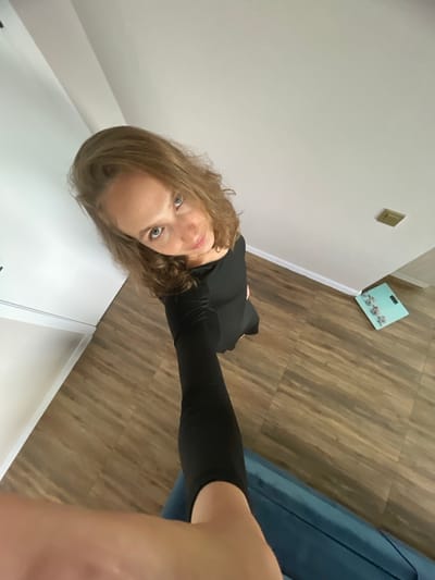 This is a selfie taken by a person with shoulder-length brown hair and blue eyes. They are wearing a black long-sleeved shirt and black leggings. The person is standing indoors, and the scene includes a white wall, light brown wooden flooring, and a blue couch or chair. In the background, there is a light blue scale with a floral pattern on it. A light switch is visible on the white wall. The lighting suggests it is daytime. The person is looking directly at the camera with a neutral expression. The overall setting appears to be a residential interior, possibly a bedroom or hallway.