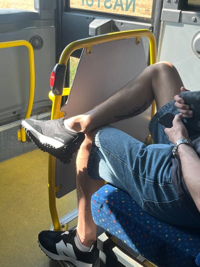 The image shows a person sitting on a bus. The person is wearing denim shorts and black and white sneakers. The bus interior features blue patterned seats and yellow handrails. A red "STOP" button is visible on a black panel. Sunlight streams in, suggesting it is daytime. The person is holding a phone in their left hand and a wallet or small object in their right. The scene is indoors, within a public transportation vehicle. Text on a sign visible through the window reads "IVECO".