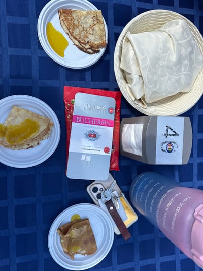 This media file captures a close-up, overhead view of a picnic spread on a blue checkered tablecloth, likely in Duvankul', Russia. The scene is set outdoors, suggested by the casual arrangement of food and the presence of a reusable water bottle.

The main subjects are several plates of crepes, some topped with a bright yellow sauce, possibly honey or jam. A decorative white basket holds folded napkins. Adjacent to the basket is a napkin dispenser with the number "40" and a colorful circular logo, indicating a celebration or milestone. A packaged chocolate bar, identified by the text "BUCHERON CHOCOLATE" and "МОЛОЧНЫЙ ШОКОЛАД С кусочками малины" (Milk chocolate with raspberry pieces), is prominently displayed.

Other objects include a smartphone, keys with a leather keychain, and a large pink and blue water bottle marked with encouraging hourly messages like "GOOD MORNING," "HYDRATE YOURSELF," and "YOU SUCCEEDED," suggesting a focus on well-being.

The overall mood is relaxed and celebratory, with the food and decorative elements hinting at a pleasant outdoor gathering. The time of day is not explicitly stated, but the well-lit scene suggests daytime, possibly mid-morning or afternoon, given the picnic context. There are no visible people in the frame.