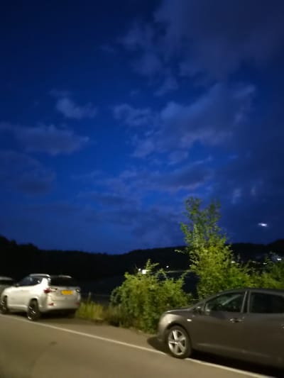 The image captures an outdoor scene at dusk or night, with a dark blue sky filled with clouds. In the foreground, a paved road runs diagonally from the bottom left to the bottom right. On the left side of the road, a white SUV is parked, facing right. Its license plate is visible and appears to be yellow. Further down the road, a dark gray sedan is parked, facing left, with its front wheel clearly visible.
Behind the road and cars, there is a body of water, possibly a lake or river, bordered by a dark, treed hillside. Dense green bushes and trees are located between the road and the water, obscuring the view of the shoreline. The foliage is illuminated, suggesting artificial lighting. The overall atmosphere is dimly lit, consistent with evening or night conditions. The location is identified as Differdange, Luxembourg.